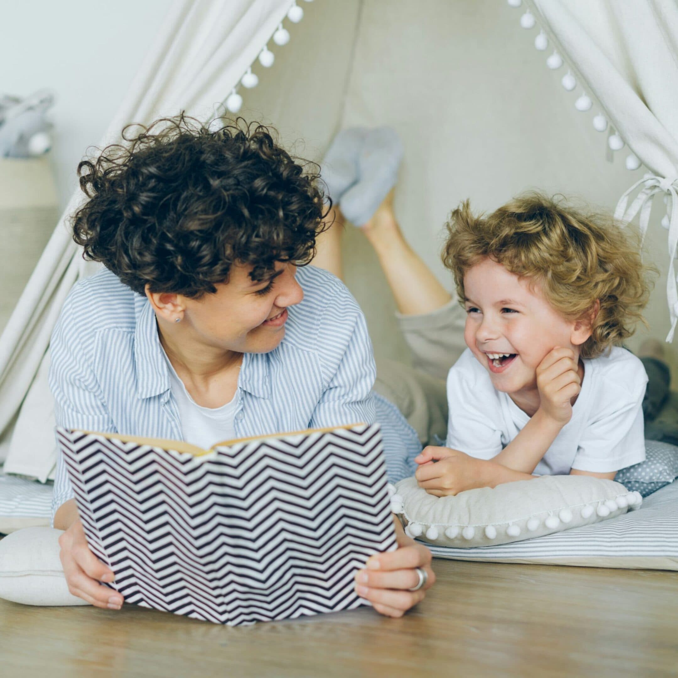 Parent Coaching An adult and a child with curly hair smile at each other while lying on the floor inside a cozy play tent. The adult holds an open book, and the child rests on a pillow, both looking happy and engaged.