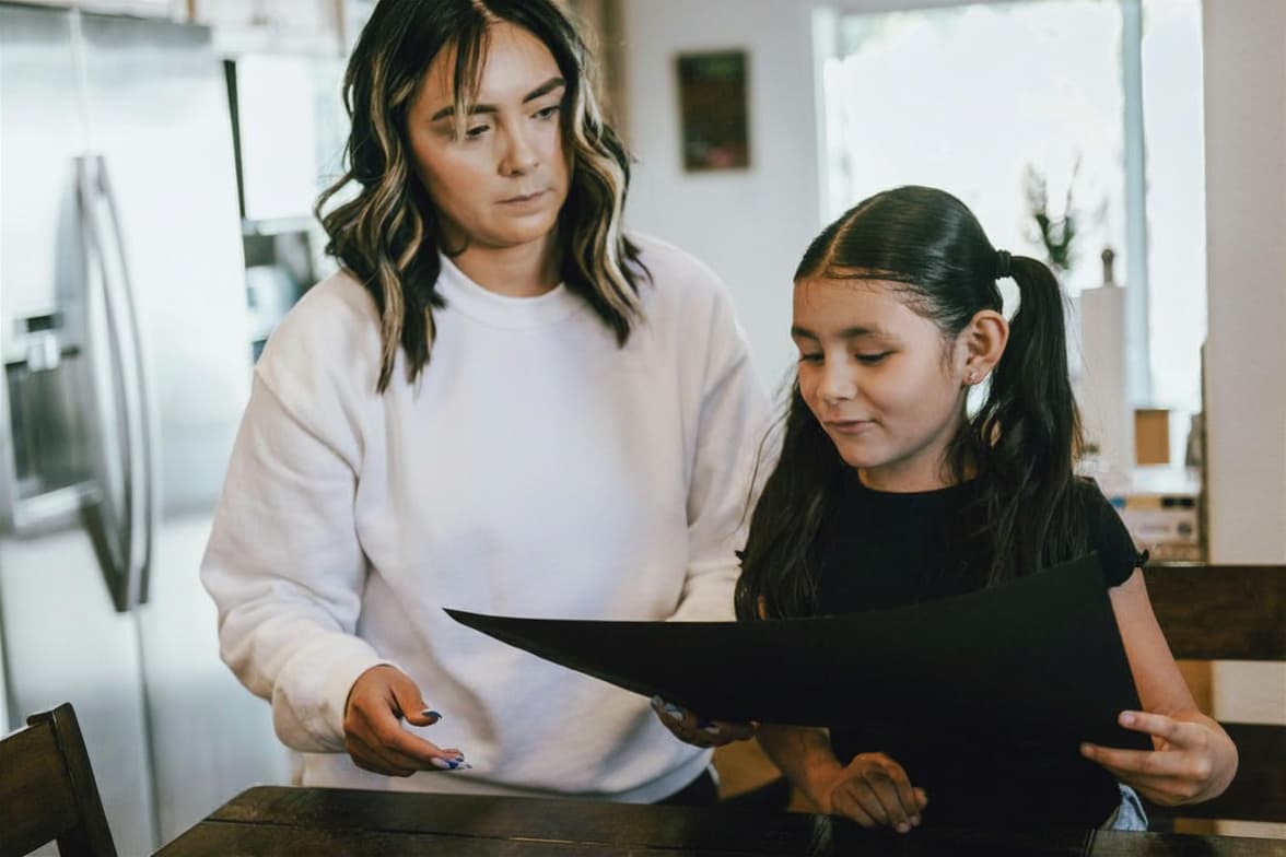 A woman and a young girl stand together indoors, looking at a large black sheet of paper on a table. The woman appears to be helping or instructing the girl. The setting looks like a kitchen or dining area.