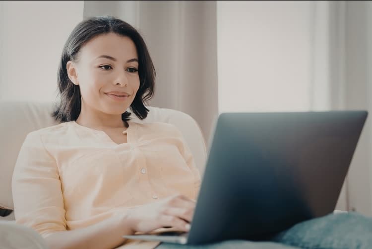 A woman sits comfortably on a couch, smiling as she works on a laptop. She wears a light-colored blouse and appears relaxed in a bright, cozy room with neutral-toned curtains in the background.