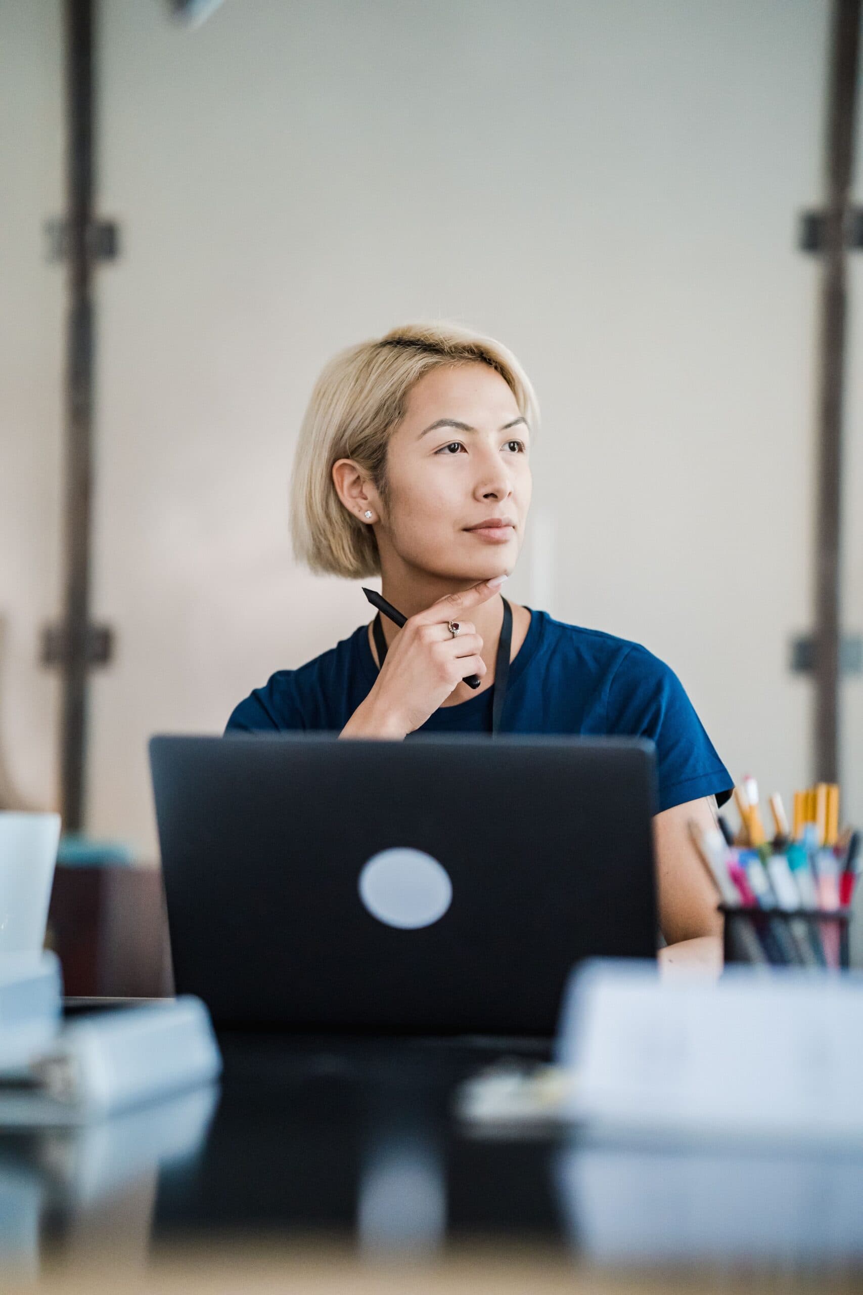 A person with short blonde hair sits at a desk with a laptop, pens, and papers, looking thoughtfully to the side, with one hand resting on their chin.