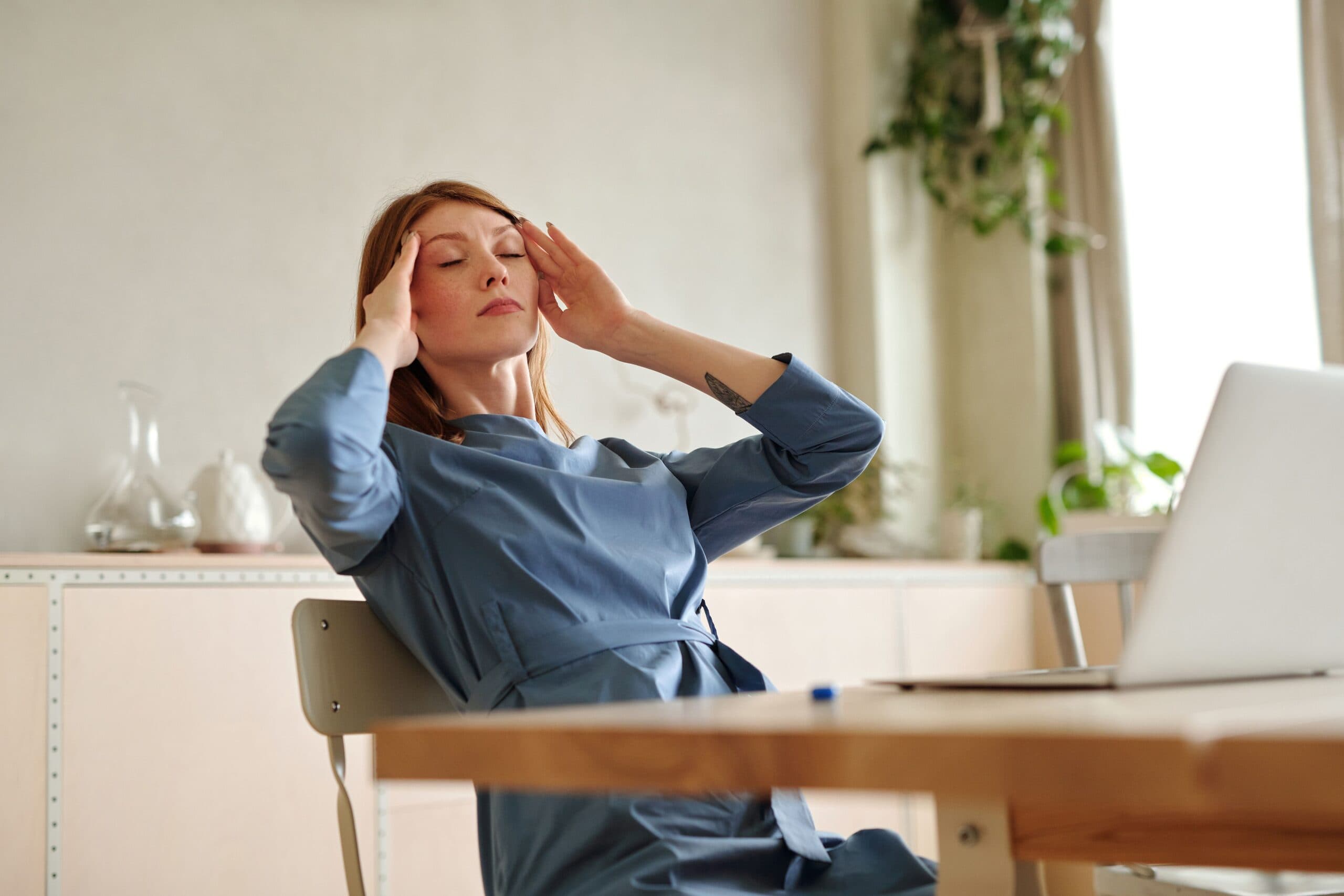Woman in a blue dress sits at a desk with her eyes closed, touching her temples as if she has a headache or is stressed, with a laptop and plants in the background.