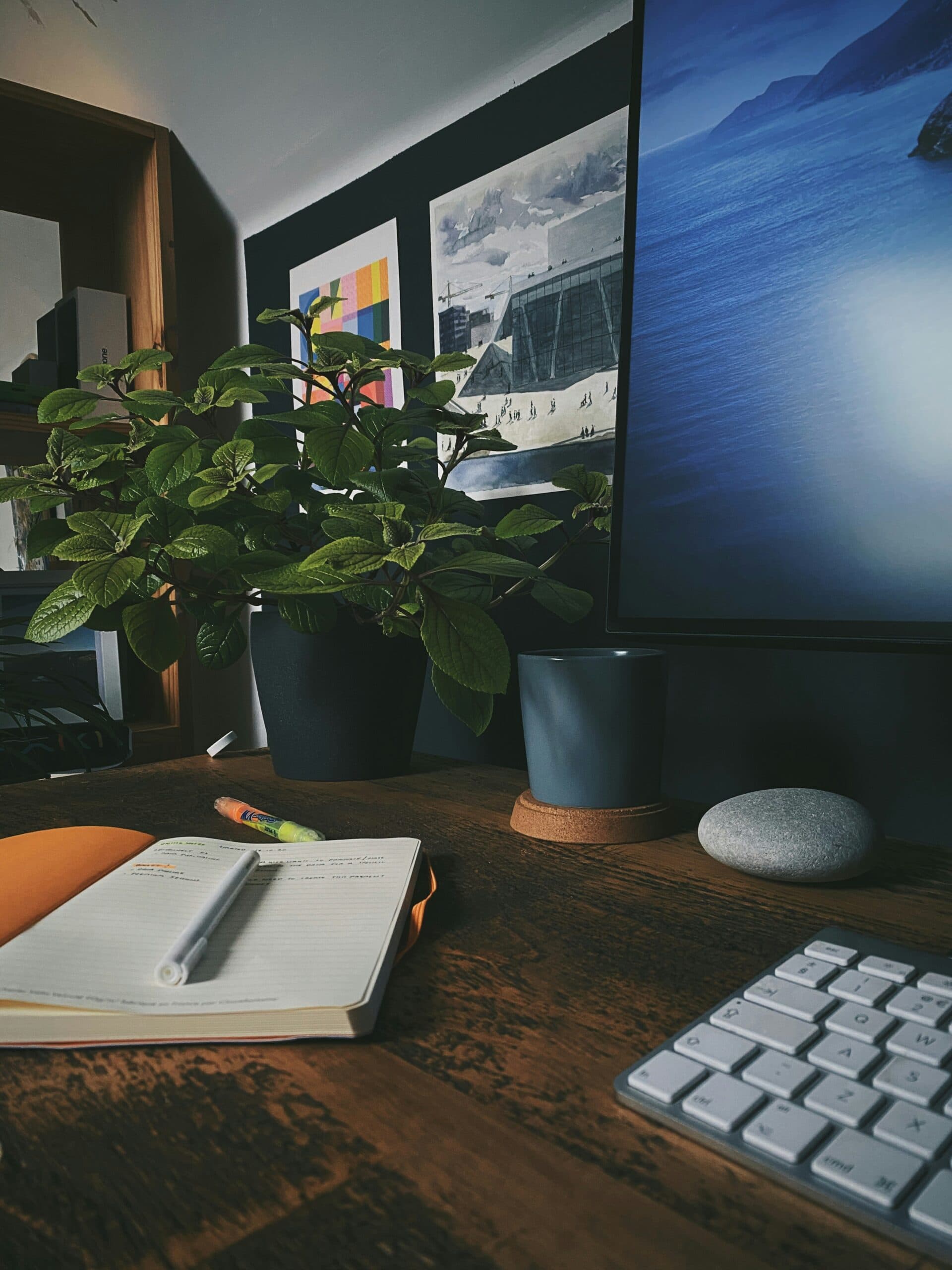 A desk with a keyboard, open notebook, pen, jar of crayons, potted plant, a mug, and part of a computer monitor displaying a seascape. Wall art and shelves are in the background.