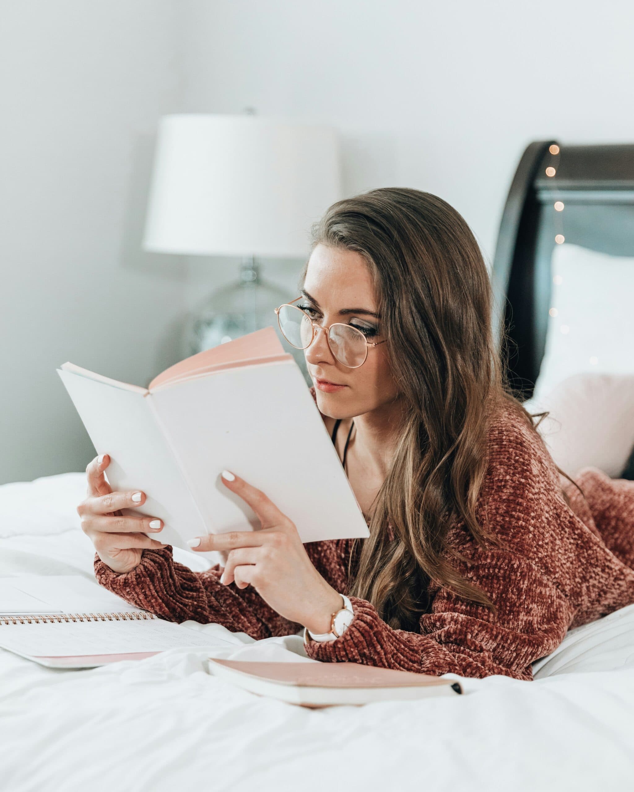 A woman with long brown hair and glasses lies on a bed, reading a book. She wears a cozy maroon sweater, with notebooks beside her. A lamp and headboard are visible in the softly lit bedroom.