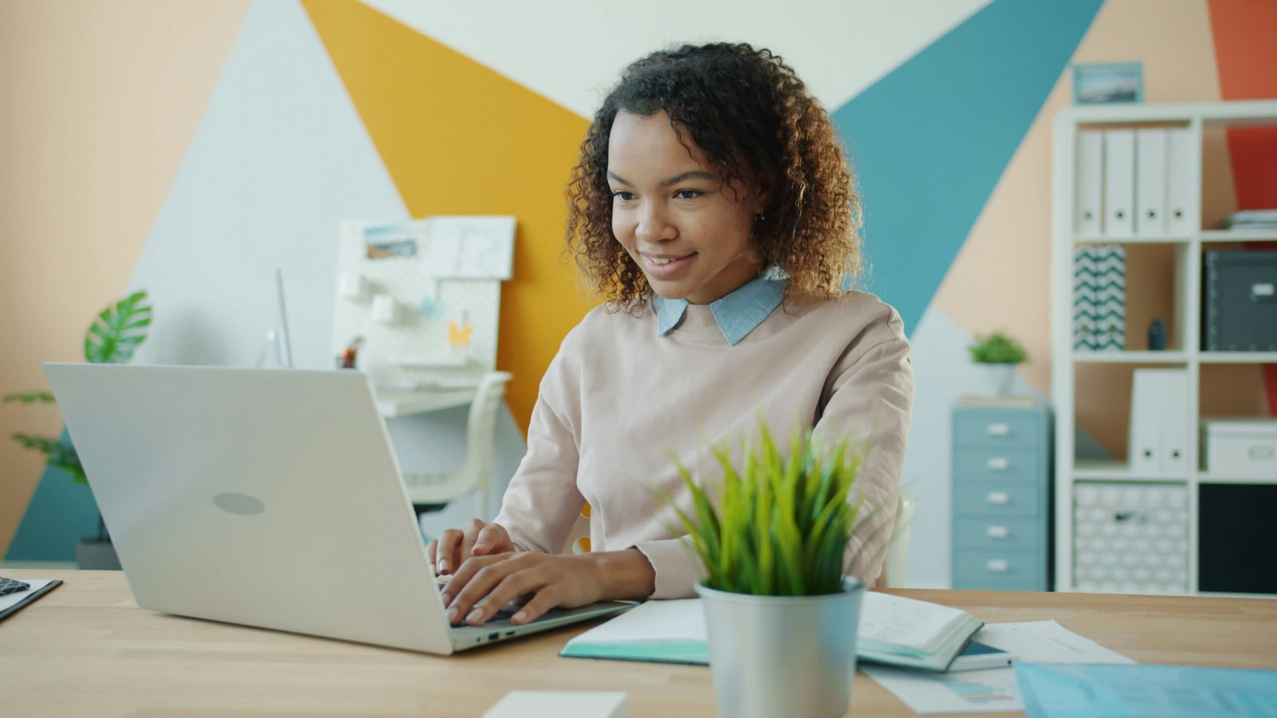 A woman sits at a desk using a laptop, smiling, with a green plant and notebooks nearby. The background features a colorful geometric wall and office shelves.