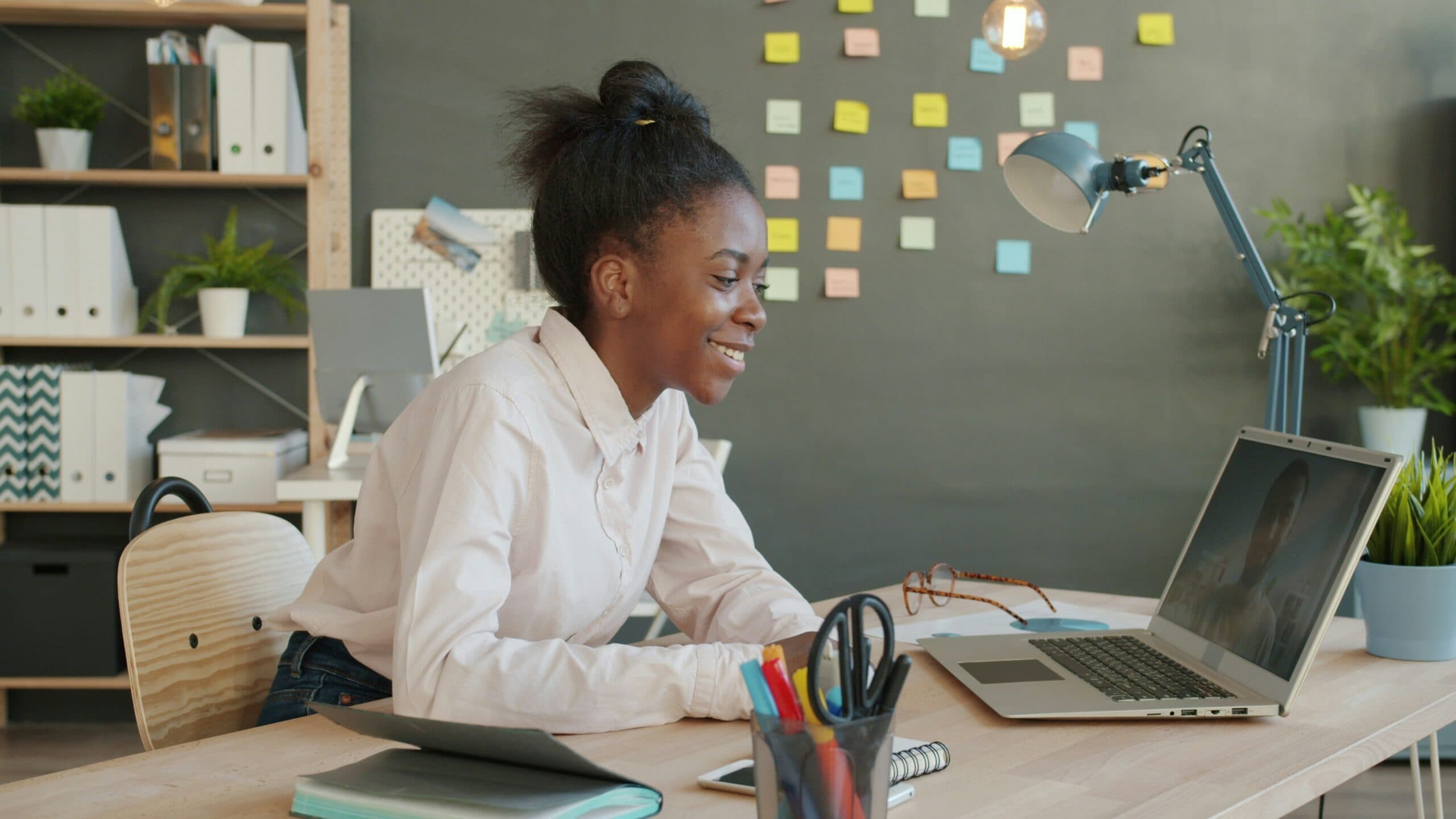 A woman sits at a desk smiling at her laptop. The background has colorful sticky notes on a chalkboard wall, office supplies, a lamp, and plants.